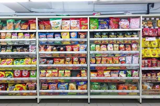 A supermarket aisle filled with a variety of colorful packaged snacks arranged on metal shelving units. Different brands and flavors of chips and other snacks are prominently displayed, creating a vibrant and diverse display of products.