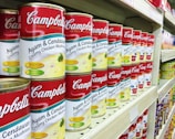 A supermarket shelf filled with rows of Campbell's canned soups, specifically labeled as 'Ayam & Cendawan Creamy Chicken Mushroom.' The cans feature a vibrant red and white label with images of creamy soup and green herbs. The shelf is well-organized, showing multiple stacks of identical cans, creating a sense of abundance and variety.