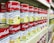A supermarket shelf filled with rows of Campbell's canned soups, specifically labeled as 'Ayam & Cendawan Creamy Chicken Mushroom.' The cans feature a vibrant red and white label with images of creamy soup and green herbs. The shelf is well-organized, showing multiple stacks of identical cans, creating a sense of abundance and variety.