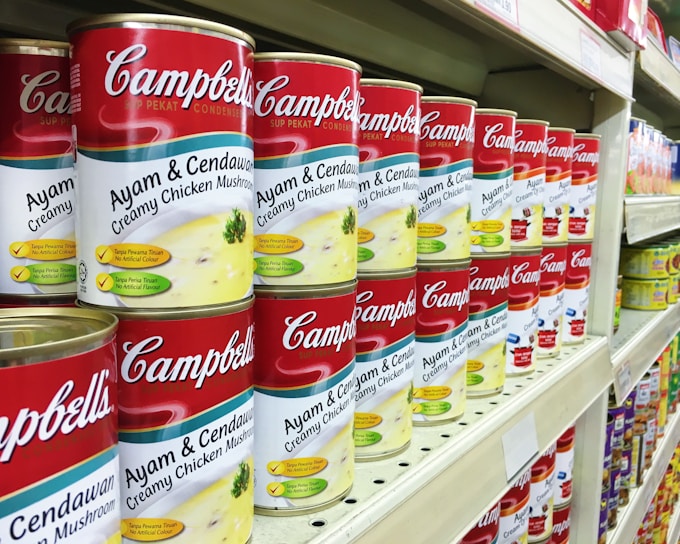 A supermarket shelf filled with rows of Campbell's canned soups, specifically labeled as 'Ayam & Cendawan Creamy Chicken Mushroom.' The cans feature a vibrant red and white label with images of creamy soup and green herbs. The shelf is well-organized, showing multiple stacks of identical cans, creating a sense of abundance and variety.
