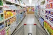 A grocery store aisle displays neatly organized shelves filled with various packaged goods and household products, including tissues, toilet paper, and cleaning supplies. A shopping cart is positioned in the foreground, facing down the aisle under bright fluorescent lighting.