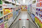 A grocery store aisle displays neatly organized shelves filled with various packaged goods and household products, including tissues, toilet paper, and cleaning supplies. A shopping cart is positioned in the foreground, facing down the aisle under bright fluorescent lighting.