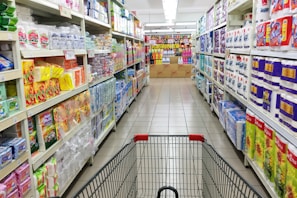 A grocery store aisle displays neatly organized shelves filled with various packaged goods and household products, including tissues, toilet paper, and cleaning supplies. A shopping cart is positioned in the foreground, facing down the aisle under bright fluorescent lighting.