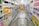 A grocery store aisle displays neatly organized shelves filled with various packaged goods and household products, including tissues, toilet paper, and cleaning supplies. A shopping cart is positioned in the foreground, facing down the aisle under bright fluorescent lighting.