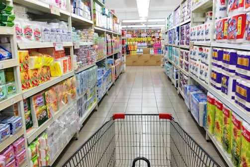 A grocery store aisle displays neatly organized shelves filled with various packaged goods and household products, including tissues, toilet paper, and cleaning supplies. A shopping cart is positioned in the foreground, facing down the aisle under bright fluorescent lighting.