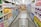A grocery store aisle displays neatly organized shelves filled with various packaged goods and household products, including tissues, toilet paper, and cleaning supplies. A shopping cart is positioned in the foreground, facing down the aisle under bright fluorescent lighting.