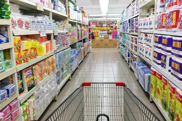 A grocery store aisle displays neatly organized shelves filled with various packaged goods and household products, including tissues, toilet paper, and cleaning supplies. A shopping cart is positioned in the foreground, facing down the aisle under bright fluorescent lighting.