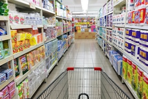 A grocery store aisle displays neatly organized shelves filled with various packaged goods and household products, including tissues, toilet paper, and cleaning supplies. A shopping cart is positioned in the foreground, facing down the aisle under bright fluorescent lighting.