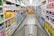 A grocery store aisle displays neatly organized shelves filled with various packaged goods and household products, including tissues, toilet paper, and cleaning supplies. A shopping cart is positioned in the foreground, facing down the aisle under bright fluorescent lighting.