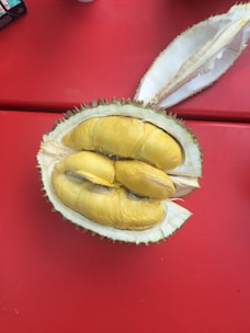 A cut-open durian fruit displaying its creamy yellow flesh sits on a red surface. The outer shell of the durian is green and thorny, with some of the spiky exterior visible. The pulp segments inside are clearly visible, showing the distinct texture and color of the fruit.