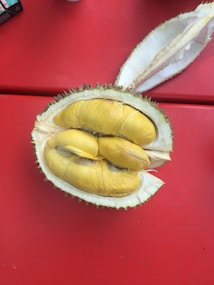 A cut-open durian fruit displaying its creamy yellow flesh sits on a red surface. The outer shell of the durian is green and thorny, with some of the spiky exterior visible. The pulp segments inside are clearly visible, showing the distinct texture and color of the fruit.