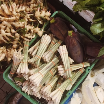 A selection of fresh produce including ginger, lemongrass bundles, banana flowers, and leafy greens neatly arranged in crates at a market.