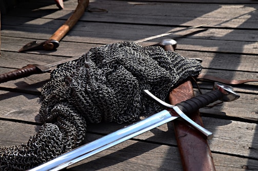 A pile of chainmail lies on a wooden plank floor alongside several medieval weapons, including a sword with a crossguard and a leather scabbard. The setting appears rustic and possibly outdoor, with shadows casting across the scene.