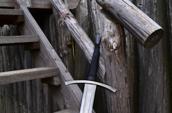 A wooden staircase leads up beside a rustic wooden fence. A large medieval-style sword rests against one of the wooden posts, featuring a broad blade and a cross-shaped hilt.