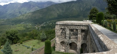 A historic stone structure with arched openings is situated amidst a lush, green landscape. The foreground includes well-maintained gardens with neatly trimmed bushes and trees. Beyond the garden, a vast expanse of dense forest extends to the foot of distant hills and mountains, under a sky partially filled with clouds.