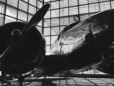 A vintage airplane is housed in a large building with a grid-like window structure. The light casts intricate reflections and shadows on the airplane's metallic body, highlighting its riveted panels and the smooth curve of its propeller.