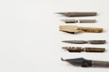 Several metallic calligraphy pen nibs are arranged in a diagonal line on a plain white surface. The nibs vary in shape and size, with some having decorative engravings and others appearing more functional.
