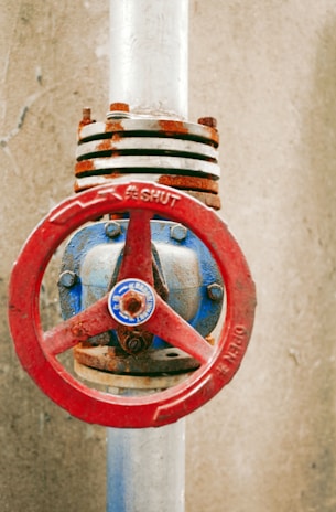 A close-up view of a rusted valve attached to a vertical pipe against a concrete background. The valve has a red circular handle with the word 'SHUT' embossed on it, while the pipe is silver and the valve body is painted blue with rust showing through.