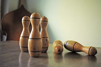 A set of wooden bowling pins are arranged on a wooden surface, with four standing upright and two lying on their sides. The lighting casts soft shadows, giving the scene a warm, rustic feel.