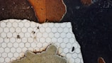 A close-up of a surface featuring white hexagonal tiles laid on a contrasting dark and cracked background. The tiles appear aged and stained, with visible dirt and imperfections. The surrounding surface shows patches of worn texture and peeling, reddish-brown paint, suggesting decay and age.