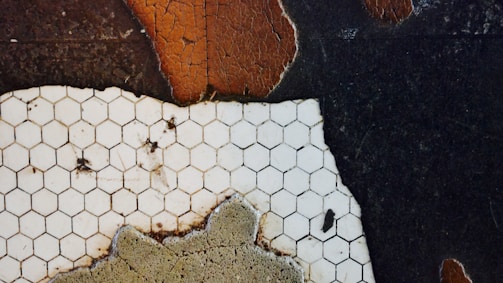 A close-up of a surface featuring white hexagonal tiles laid on a contrasting dark and cracked background. The tiles appear aged and stained, with visible dirt and imperfections. The surrounding surface shows patches of worn texture and peeling, reddish-brown paint, suggesting decay and age.