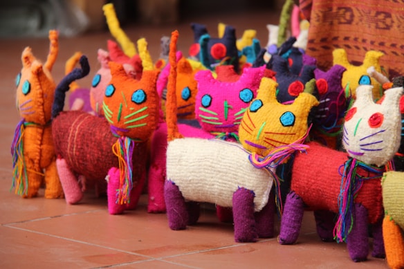 The image features a collection of colorful, handmade cat dolls arranged in close proximity. These dolls are made using a variety of vibrant fabrics and threads. Each cat has a whimsical design with bright blue eyes, whiskers, and a colorful scarf around its neck. The setting seems to be indoors with a tiled floor.