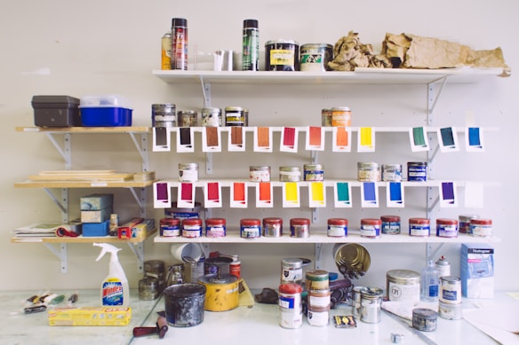 A wall-mounted shelving unit displays an assortment of paint cans, bottles, and spray paint. Swatches of various paint colors hang below some shelves for display. Other items, like a Windex bottle and plastic wrap, are placed on the table. The shelves are filled with art supplies, and the area is organized with an artistic feel.