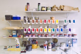 A wall-mounted shelving unit displays an assortment of paint cans, bottles, and spray paint. Swatches of various paint colors hang below some shelves for display. Other items, like a Windex bottle and plastic wrap, are placed on the table. The shelves are filled with art supplies, and the area is organized with an artistic feel.