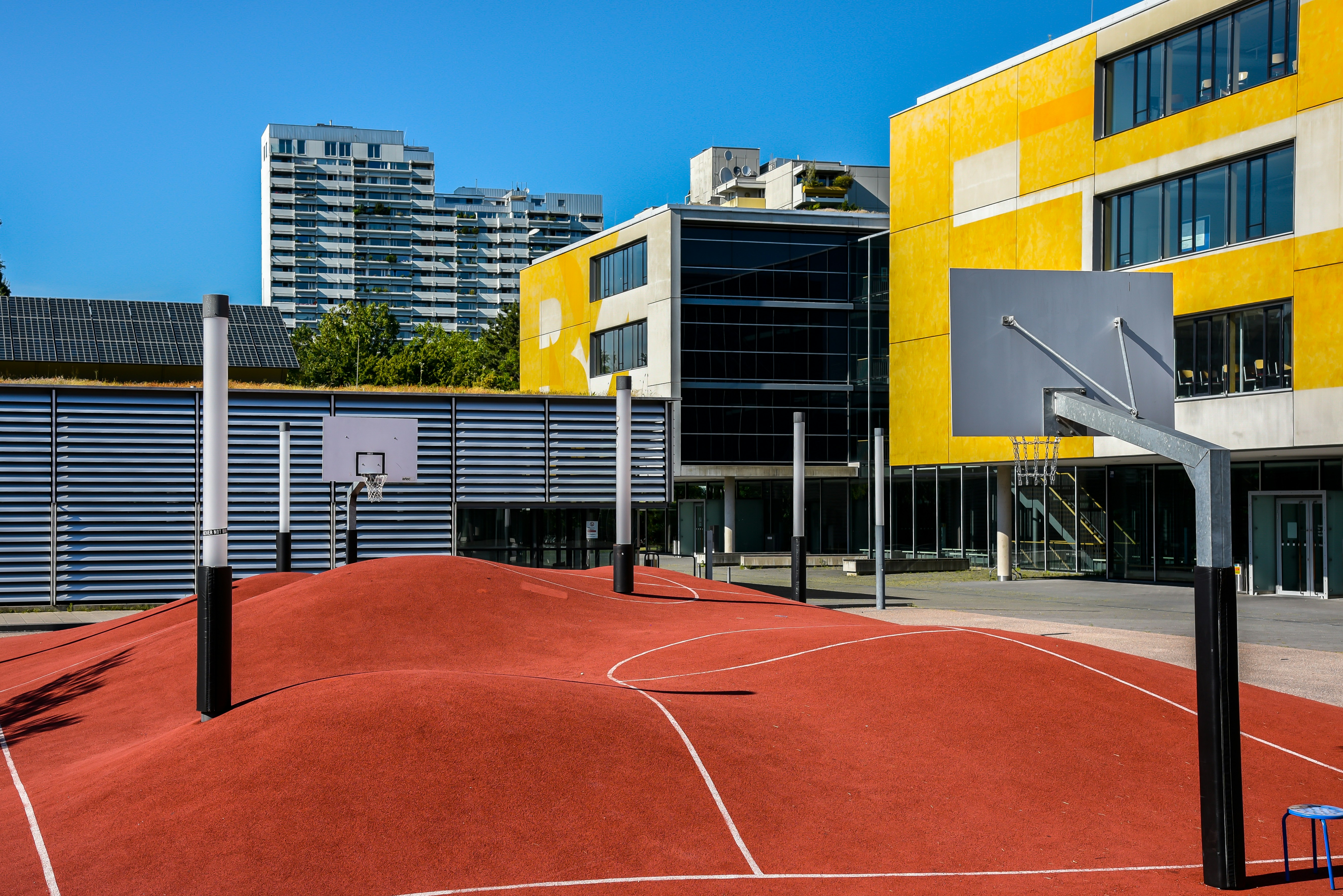 A modern urban basketball court features an undulating red surface with marked lines, surrounded by several basketball hoops with backboards. Tall residential buildings and a yellow-accented modern building serve as the backdrop under a clear blue sky.