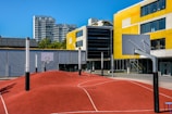 A modern urban basketball court features an undulating red surface with marked lines, surrounded by several basketball hoops with backboards. Tall residential buildings and a yellow-accented modern building serve as the backdrop under a clear blue sky.