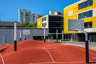 A modern urban basketball court features an undulating red surface with marked lines, surrounded by several basketball hoops with backboards. Tall residential buildings and a yellow-accented modern building serve as the backdrop under a clear blue sky.