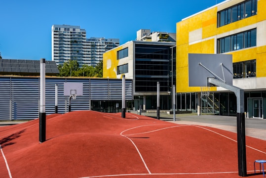 A modern urban basketball court features an undulating red surface with marked lines, surrounded by several basketball hoops with backboards. Tall residential buildings and a yellow-accented modern building serve as the backdrop under a clear blue sky.