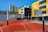 A modern urban basketball court features an undulating red surface with marked lines, surrounded by several basketball hoops with backboards. Tall residential buildings and a yellow-accented modern building serve as the backdrop under a clear blue sky.