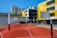A modern urban basketball court features an undulating red surface with marked lines, surrounded by several basketball hoops with backboards. Tall residential buildings and a yellow-accented modern building serve as the backdrop under a clear blue sky.