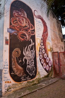 A vibrant mural on a wall featuring detailed illustrations of octopus tentacles. The composition includes a dark background with contrasting red and white tentacles curling in various directions. The artwork incorporates handwritten text around and within the images.