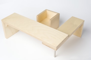 A minimalist wooden structure featuring a U-shaped design with a flat surface and a square compartment, crafted from light-colored plywood.