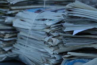 A collection of stacked newspapers bound together with string. The papers are piled in a somewhat haphazard manner, with edges jutting out in various directions. The color scheme is predominantly shades of gray and blue, suggesting the papers may have aged or been stored for a while.