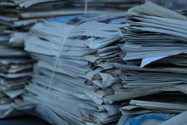 A collection of stacked newspapers bound together with string. The papers are piled in a somewhat haphazard manner, with edges jutting out in various directions. The color scheme is predominantly shades of gray and blue, suggesting the papers may have aged or been stored for a while.
