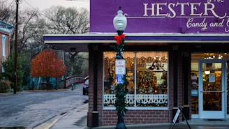 A quaint candy and gift shop adorned with festive decorations. The storefront has a bright purple awning and holiday wreaths, with a vintage street lamp decorated for the season. Inside the windows, warm lights and candy displays create a welcoming atmosphere. A no parking bus stop sign is visible on the street corner.