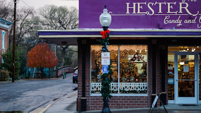 A quaint candy and gift shop adorned with festive decorations. The storefront has a bright purple awning and holiday wreaths, with a vintage street lamp decorated for the season. Inside the windows, warm lights and candy displays create a welcoming atmosphere. A no parking bus stop sign is visible on the street corner.