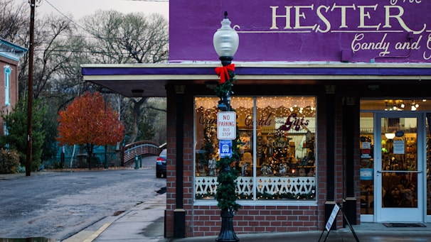 A quaint candy and gift shop adorned with festive decorations. The storefront has a bright purple awning and holiday wreaths, with a vintage street lamp decorated for the season. Inside the windows, warm lights and candy displays create a welcoming atmosphere. A no parking bus stop sign is visible on the street corner.
