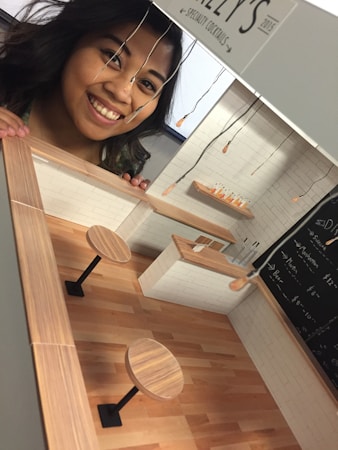 A woman smiling next to a miniature model of a bar or cafe. The model features wooden tables, a bar counter, and hanging light fixtures. The walls of the model are white with a blackboard menu on one side.