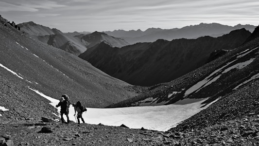 Two hikers with backpacks and trekking poles are climbing a rocky mountain slope. Snow patches are visible on the ground, and rugged mountain peaks stretch across the horizon under a clear sky.