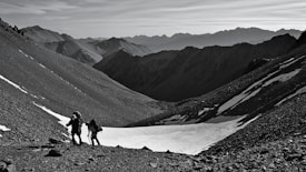 Two hikers with backpacks and trekking poles are climbing a rocky mountain slope. Snow patches are visible on the ground, and rugged mountain peaks stretch across the horizon under a clear sky.