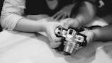 Hands of two individuals handling a vintage-style camera, focusing on the camera’s top control dials and lens. The image is in black and white, with a soft focus on the hands and camera, creating an intimate and nostalgic feel.