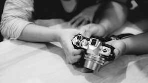 Hands of two individuals handling a vintage-style camera, focusing on the camera’s top control dials and lens. The image is in black and white, with a soft focus on the hands and camera, creating an intimate and nostalgic feel.