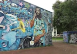 A large, vibrant mural located on a building wall, depicting a DJ with dreadlocks mixing a vinyl record on a turntable. The DJ is surrounded by graffiti art, colorful musical elements, such as a trumpet, speakers, sound equipment, and abstract designs. The background contains multicolored graffiti text and stars, adding to the lively urban atmosphere. Trees and other graffiti-covered structures can also be seen nearby.