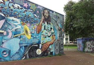 A large, vibrant mural located on a building wall, depicting a DJ with dreadlocks mixing a vinyl record on a turntable. The DJ is surrounded by graffiti art, colorful musical elements, such as a trumpet, speakers, sound equipment, and abstract designs. The background contains multicolored graffiti text and stars, adding to the lively urban atmosphere. Trees and other graffiti-covered structures can also be seen nearby.