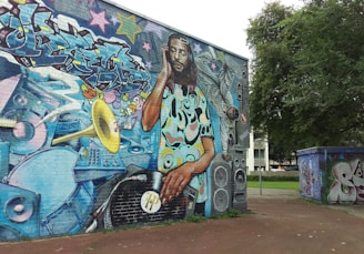 A large, vibrant mural located on a building wall, depicting a DJ with dreadlocks mixing a vinyl record on a turntable. The DJ is surrounded by graffiti art, colorful musical elements, such as a trumpet, speakers, sound equipment, and abstract designs. The background contains multicolored graffiti text and stars, adding to the lively urban atmosphere. Trees and other graffiti-covered structures can also be seen nearby.