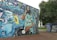 A large, vibrant mural located on a building wall, depicting a DJ with dreadlocks mixing a vinyl record on a turntable. The DJ is surrounded by graffiti art, colorful musical elements, such as a trumpet, speakers, sound equipment, and abstract designs. The background contains multicolored graffiti text and stars, adding to the lively urban atmosphere. Trees and other graffiti-covered structures can also be seen nearby.